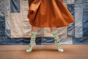 woman standing on her tip toes  in front of a patchwork denim background wearing a rust colored dress and minty green, patterned organic cotton socks